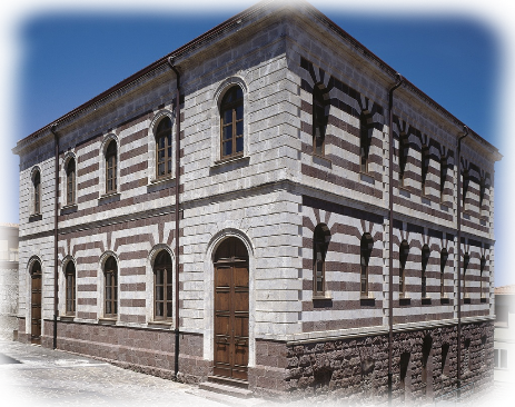 Villanova Monteleone e il Palatu 'e sas Iscolas fu realizzato tra il 1890 e il 1920 su progetto dell'ingegnere Achille Dettori. La singolare costruzione ha forme solide e compatte e, sfruttando il dislivello del terreno, passa da due a tre piani, sottolineando il basamento con un alto zoccolo di trachite rossa. Fasce bicrome rosse e grigie caratterizzano fortemente l'edificio, ricorrendo ad un linguaggio neomedioevale, che richiama la bicromia dell'architettura romanica in Sardegna. Le finestre si sviluppano regolarmente e simmetricamente ed hanno tutte forma ad arco come i portali d'ingresso. Datazione soggetto: 1890-1920.